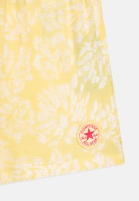 Tissu jaune avec un motif floral, comportant un patch circulaire rouge portant l'inscription "CONVERSE ALL STAR" et des détails de surface texturée.