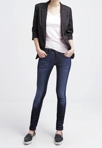 Mörkblå skinny jeans med en slät denimtextur, bärs med en ljusrosa topp och en svart kavaj, ihop med tofflor.