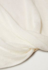 Kermanvärinen neulekangas, jossa on pehmeä rakenne ja kierretty muotoilu. Materiaali näyttää kevyeltä ja löysästi kudotulta.