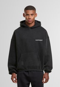 Giovane uomo che indossa un hoodie nero con il testo "SundayRunningClub" sul petto e pantaloni neri, in piedi davanti a uno sfondo grigio uniforme.