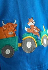 Lapsas, kas vada zaļu traktoru, kas velk piekabi ar brūnu bulli, izšūts uz zila auduma.