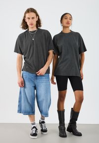 Twee modellen dragen een donkergrijze oversized T-shirt. Het ene model combineert het met lichtblauwe oversized shorts en zwarte sneakers; het andere met zwarte fietsbroek en hoge zwarte laarzen.