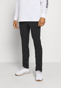 Pantalon d'extérieur noir à coupe ajustée, avec une texture lisse et des détails de couture subtils. Associé à une chemise blanche à manches longues et des baskets blanches.