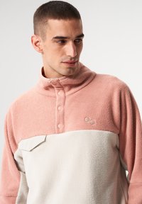 Fleece-Pullover in Hellrosa und Creme mit hohem Kragen, Knopfleiste und aufgesetzter Tasche. Seidige Textur mit gesticktem Logodetail.