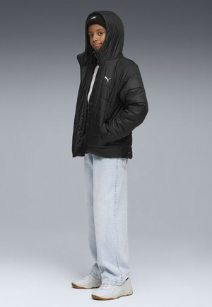Giubbotto puffer nero con cappuccio e logo bianco, abbinato a jeans baggy azzurri chiari e sneakers chiare, caratterizzati da un design e una texture semplici.