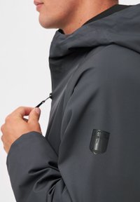 Grijze waterdichte jas met een gestructureerde capuchon, een soepele textuur en een merkpatch op de mouw. Voorzien van een verstelbaar trekkoord bij de hals.