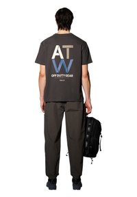 Grå kortärmad skjorta med en djärv "ATW" logotyp i kräm och blå på ryggen, kombinerad med mörka, strukturerade cargobyxor och en svart ryggsäck.