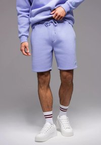 Shorts en mélange de coton lavande clair avec une taille élastique et un cordon de serrage. Associés à des baskets blanches et des chaussettes à rayures pour les chevilles.