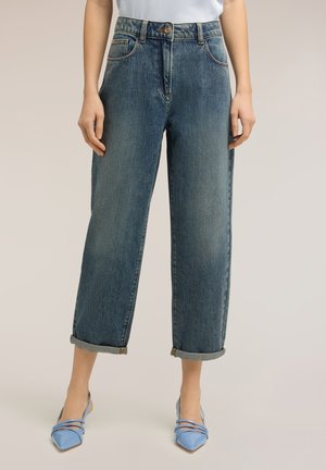 Frau trägt nach oben gerollte gerade geschnittene blaue Jeans und spitze blaue Absätze, steht vor einem neutralen Hintergrund.