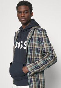 Giacca a quadri nei toni del verde, blu navy e beige, indossata sopra un hoodie blu navy con testo bianco. Il modello posa con le mani in tasca.