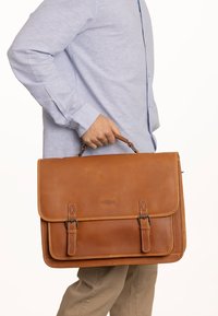 Tan læder messenger bag med en struktureret form, der har metalbeslag, dobbelte forreste spænder og en glat finish.