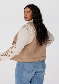 Tan mouwloos vest met een fluffy crèmekleurige kraag, gecombineerd met een gestructureerd lichtkleurig shirt met lange mouwen met geborduurde bladerenpatronen.