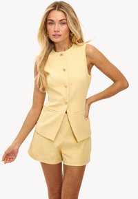 Geel mouwloos blazertje met een knoopsluiting aan de voorkant en twee zakken, gecombineerd met bijpassende gele shorts, gemaakt van een gladde stof.