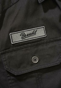 �Črna tkanina s ribjo kostjo, z sivo vezeno oznako "Brandit" in kovinskim gumbom na žepu.