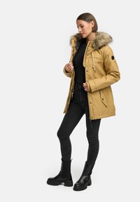 Gul parkajakke med hette i kunstpels, glidelås og trykknapper, kombinert med svarte jeans og kraftige svarte ankelstøvler.