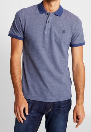 Mężczyzna ubrany w granatowo-białą, paskowaną koszulkę polo z krótkim rękawem i dżinsy na jednolitym jasnym tle.