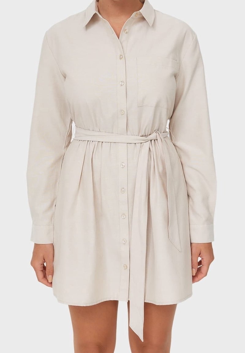Robe-chemise beige à manches longues avec boutons sur le devant, poche poitrine et ceinture nouée à la taille, portée par une personne devant un fond uni.