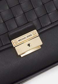 Sac en cuir noir avec une texture tissée sur le dessus et un fermoir doré gravé du nom de la marque Michael Kors.