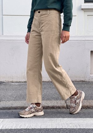 Une personne marche le long d’un mur blanc, vêtue d’un pantalon beige à jambes larges, d’un sweat-shirt vert foncé et de baskets grises.
