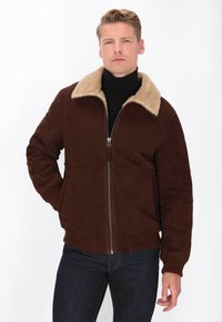 Jachetă din suede maro cu un guler din blană artificială bej, închidere cu fermoar, manșete ribbate și buzunare laterale. Purtați peste un pulover negru cu guler înalt.