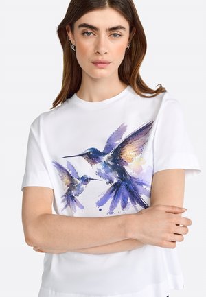 T-shirt en coton blanc présentant un motif aquarelle vibrant de deux colibris dans des teintes de violet, bleu et jaune, avec des manches courtes.