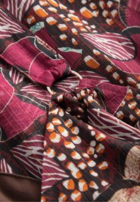 Mörkröd mönstrad tyg med orange, vit och svart motiv; har en samlad textur och en metallring som accent vid fållen.