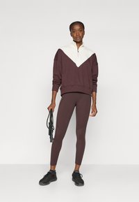 Mørkebrun zip-sweatshirt med hvit fargeblokkering; tettsittende mørkebrun leggings med buede sømmer; modellen holder en hoppsnor.