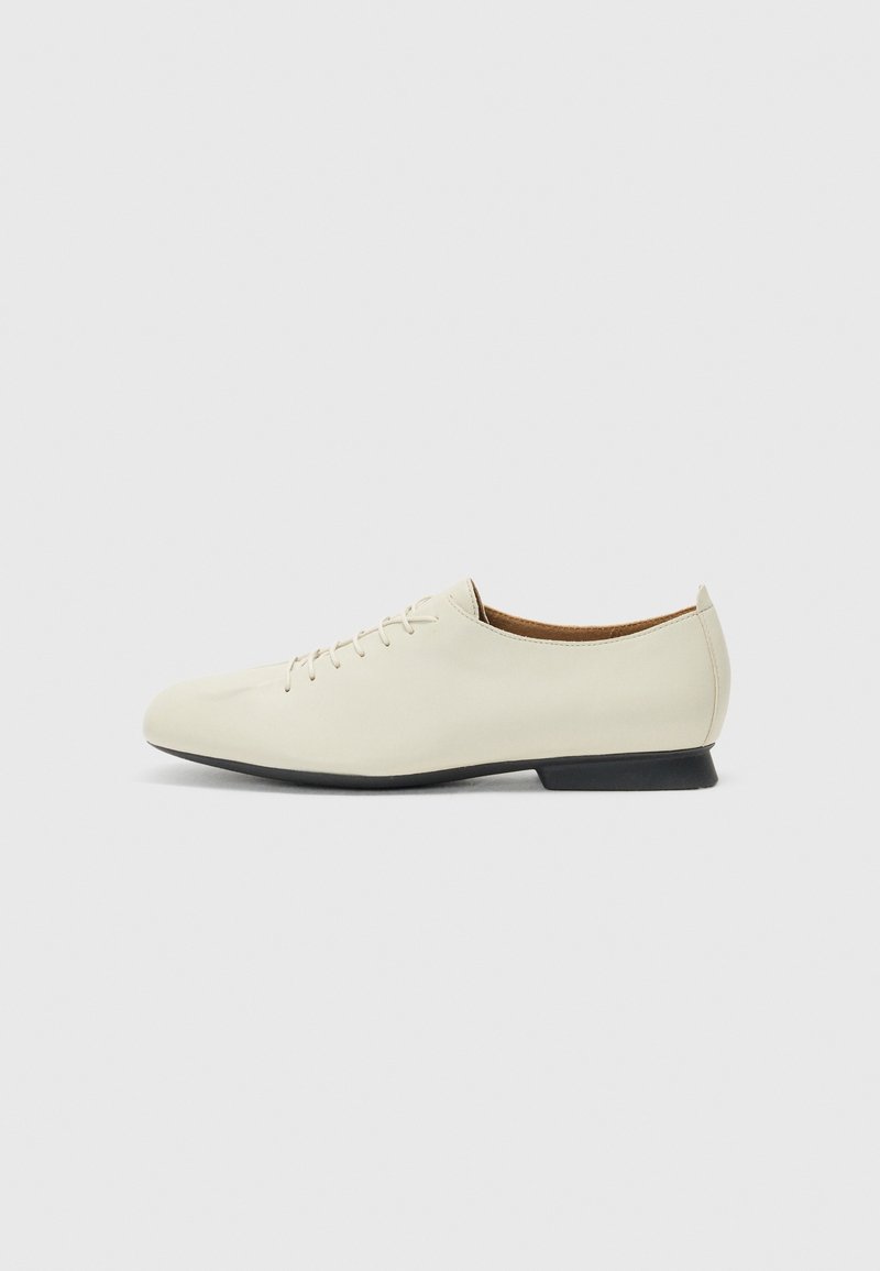 Chaussures en cuir beige au design épuré, avec un bout arrondi, un laçage minimaliste et une semelle en caoutchouc noire. Texture lisse, sans motifs visibles.