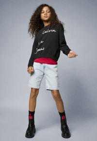 Schwarzer Pullover mit weißer Schrift, rotes T-Shirt darunter, hellblaue Denim-Shorts, schwarze Stiefeletten und schwarze Socken mit roten Akzenten.