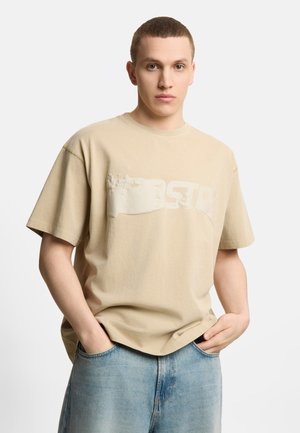 Młody mężczyzna z krótkimi włosami, ubrany w beżowy podniszczony t-shirt z logo i jasnoniebieskie jeansy, ręce w kieszeniach na jednolitym tle.