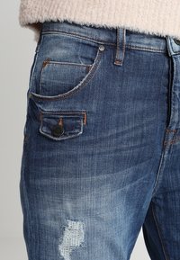 Modré denimové džíny s přiléhavým střihem, mírným vyblednutím, opotřebovanými detaily a kontrastním šitím se stříbrným knoflíkem.