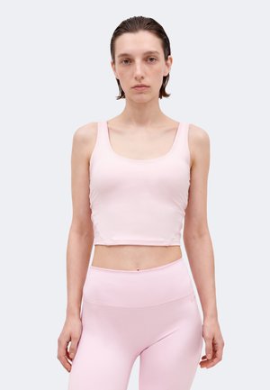 Γυναίκα με κοντά βρεγμένα μαλλιά που φοράει ανοιχτό ροζ κοντό τοπ και ψηλόμεσα κολάν μπροστά σε απλό φόντο.