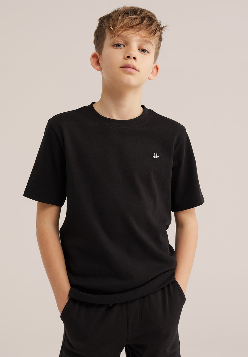 Chlapec so svetlohnedými vlasmi, oblečený v čiernom tričku s krátkym rukávom a čiernych nohaviciach, stojaci s rukami v vreckách na jednoduchom pozadí.