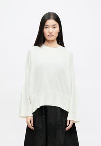 Suéter branco oversized com uma bainha canelada e mangas largas, combinado com uma saia preta com padrões florais bordados.