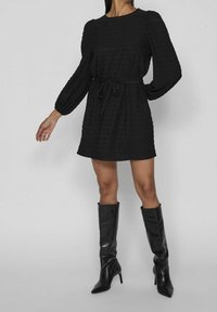 Una modelo posa con un vestido negro texturizado y un cinturón atado a la cintura, combinado con botas negras hasta la rodilla, contra un fondo gris.