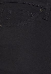 Jeans de mezclilla negra con una textura suave, que presentan un bolsillo frontal y herrajes plateados. Las costuras son sutiles y del mismo color.