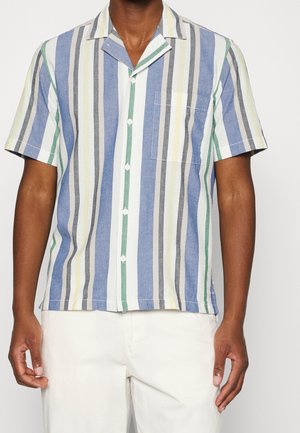 Man draagt een overhemd met korte mouwen en verticale strepen in blauw, groen, geel, grijs en wit, gecombineerd met een witte broek.