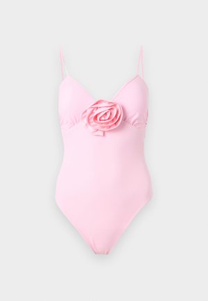 Γυναίκα στέκεται ξυπόλυτη φορώντας ροζ ολόσωμο μαγιό με λεπτές τιράντες και μεγάλο υφασμάτινο τριαντάφυλλο στο στήθος, μπροστά σε απλό ανοιχτό φόντο.