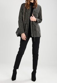 Chaqueta con patrón de camuflaje en gris oscuro, que presenta dos bolsillos en el pecho, cintura ajustada, combinada con jeans negros ajustados y botas de tobillo.
