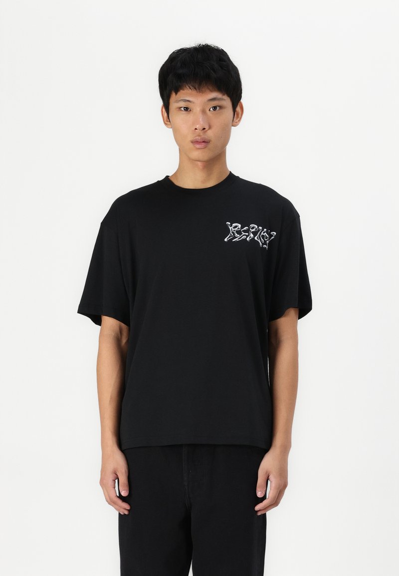 Camiseta negra de algodón con mangas cortas, de corte relajado, que presenta una impresión gráfica blanca en el lado izquierdo del pecho. Modelo vistiendo pantalones negros.