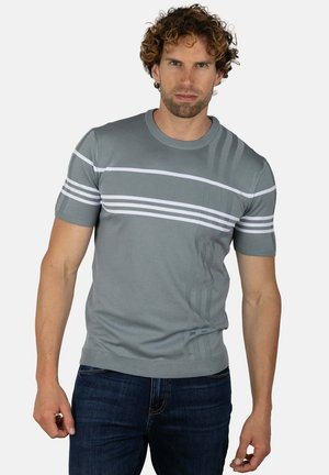 Hombre con cabello rizado que lleva un suéter gris de manga corta con rayas horizontales blancas y vaqueros azul oscuro, de pie contra un fondo blanco.