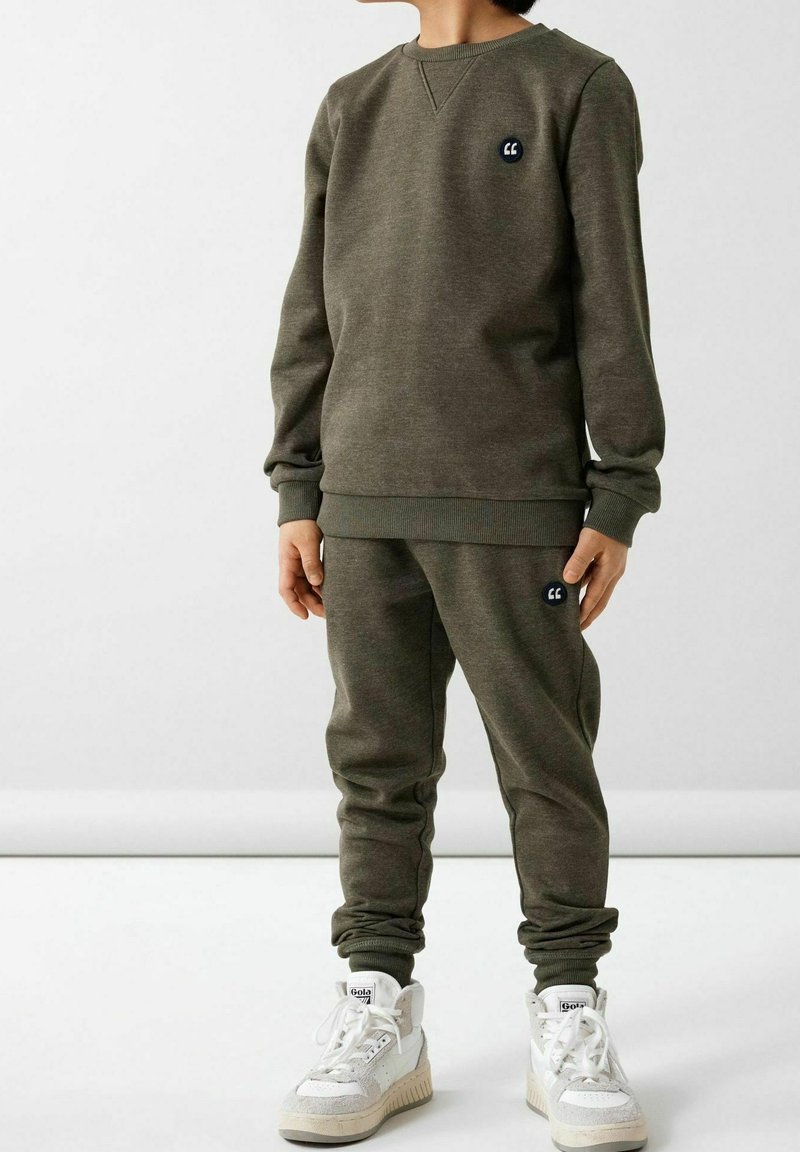 Enfant portant un sweat-shirt vert olive et un pantalon de survêtement assorti avec des baskets blanches montantes, debout devant un fond clair uni.