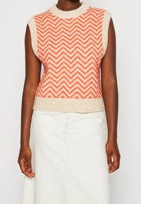 Udenærmet sweater med et rødt og cremefarvet zigzag-mønster og en beige kant. Den er lavet af struktureret stof og har et høj halsdesign.