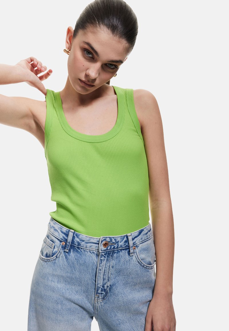 Top de tirantes verdes acanalados con tirantes anchos y escote en forma de scoop, combinado con jeans de mezclilla de tiro alto en un tono azul claro con una textura lavado.