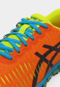 Φωτεινό πορτοκαλί αθλητικό παπούτσι με νεον κίτρινη γλώσσα και μπλε κορδόνια, με μαύρο λογότυπο και υφασμένο πάτο σε κοντινή λήψη.