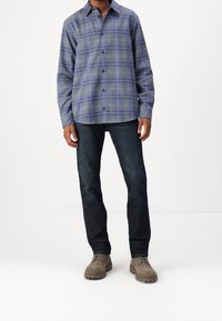 Camicia a scacchi blu e grigia con bottoni, maniche lunghe, in tessuto misto cotone, abbinata a jeans scuri in denim e scarpe marrone chiaro.