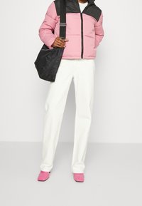 Giacca a vento con corpo rosa e spalle nere, abbinata a pantaloni dritti bianchi e scarpe rosa lucide. Porta una borsa a spalla nera.