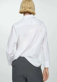 Weißes Button-up-Hemd mit langen Ärmeln, einem umgeschlagenen Kragen und einem abgerundeten Saum, kombiniert mit grauen Nadelstreifenhosen. Geschmeidige Textur.