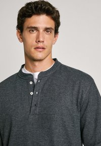 La maglietta henley a righe grigia presenta un collo alto, tre bottoni bianchi e un logo ricamato discreto. Il tessuto testurizzato aggiunge profondità.