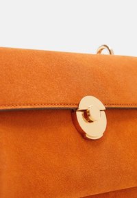 Sac à main en daim orange vif avec une fermeture circulaire dorée. Présente une texture lisse et une forme rectangulaire structurée.